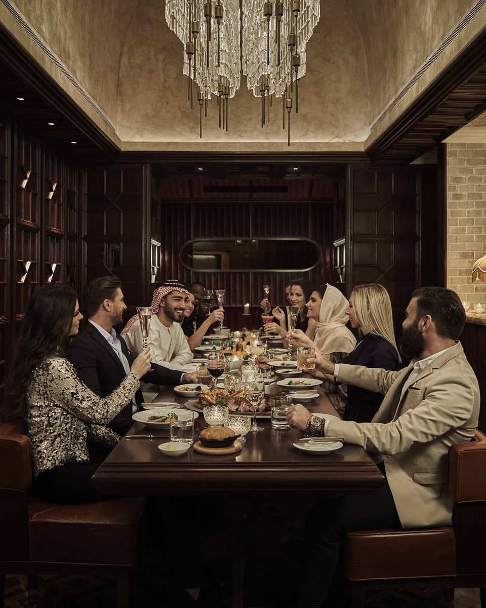 Ten people sit at a long, dark-wood rectangular table in a dark-wood panelled, speakeasy-style room with a crystal chandelier overhead and a marble bar counter and bartender to the side