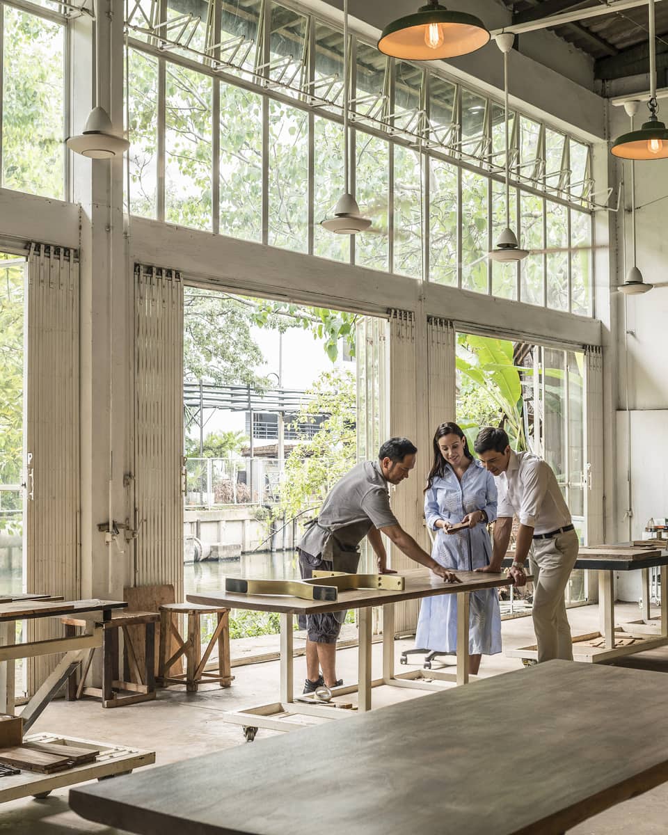 People working in a workshop with large windows, sanding and inspecting wooden furniture, while others collaborate at a table.