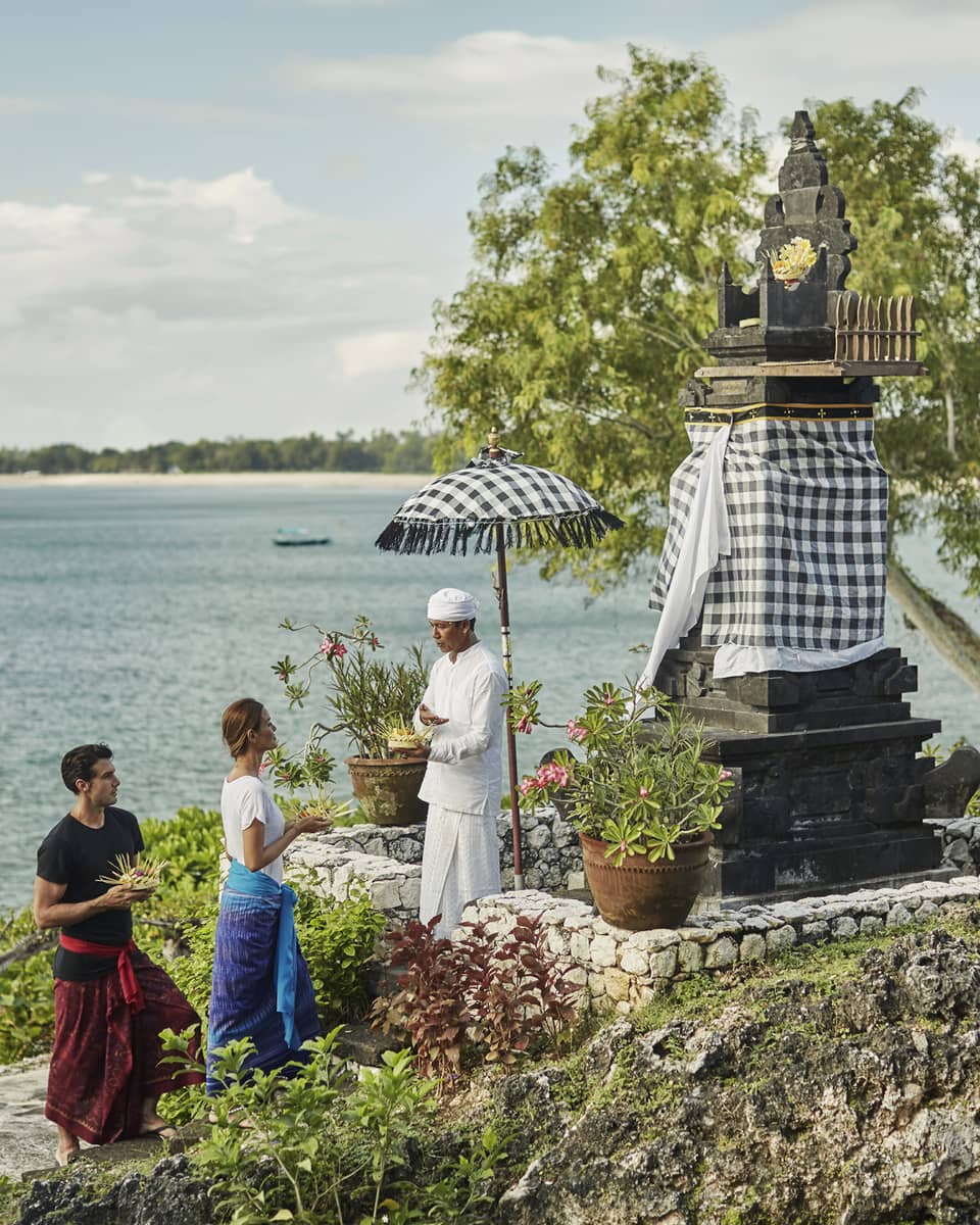 High Priest Aji Ngurah stands by shrine with black-and-white checkered umbrella, cloth, as man and woman make offering