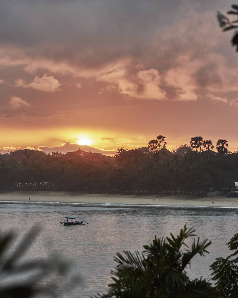 Sunrise over beach with trees and water, small boat 