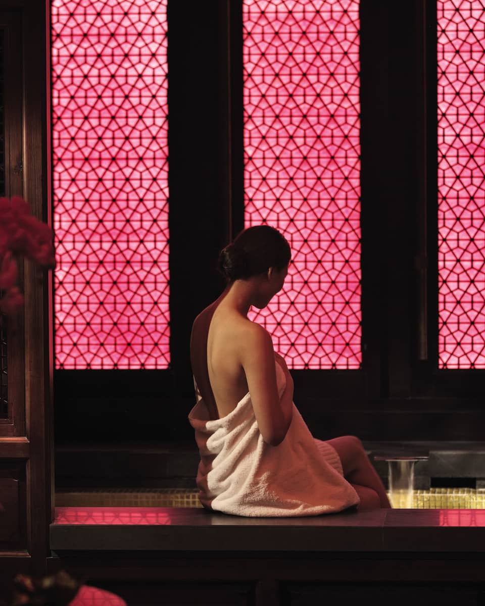 Woman wearing white towel sits on edge of dark spa tub by wall with pink lights