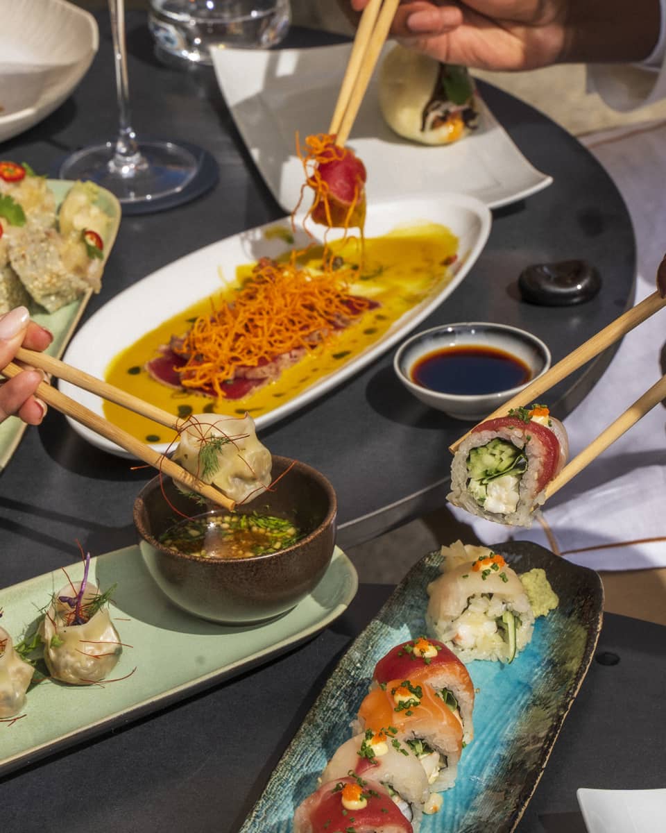 Three hands picking up sushi with chopsticks over plates of dishes
