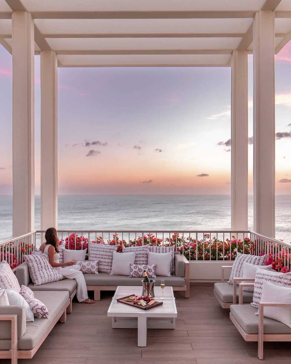 Woman sits on sofa on outdoor terrace under a white pergola overlooking the ocean