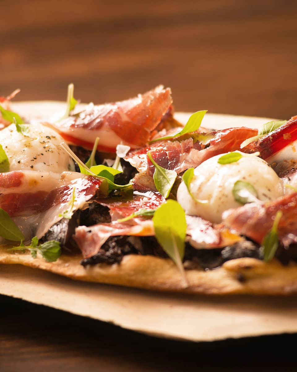 Close-up of thin crust Italian-style pizza topped with thinly-shaved cured meat, herbs, cheese