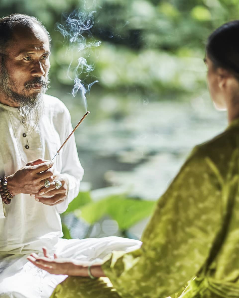 Set against a backdrop of greenery, Djik Dewa holds a stick of burning incense before a meditative cross-legged guest. 