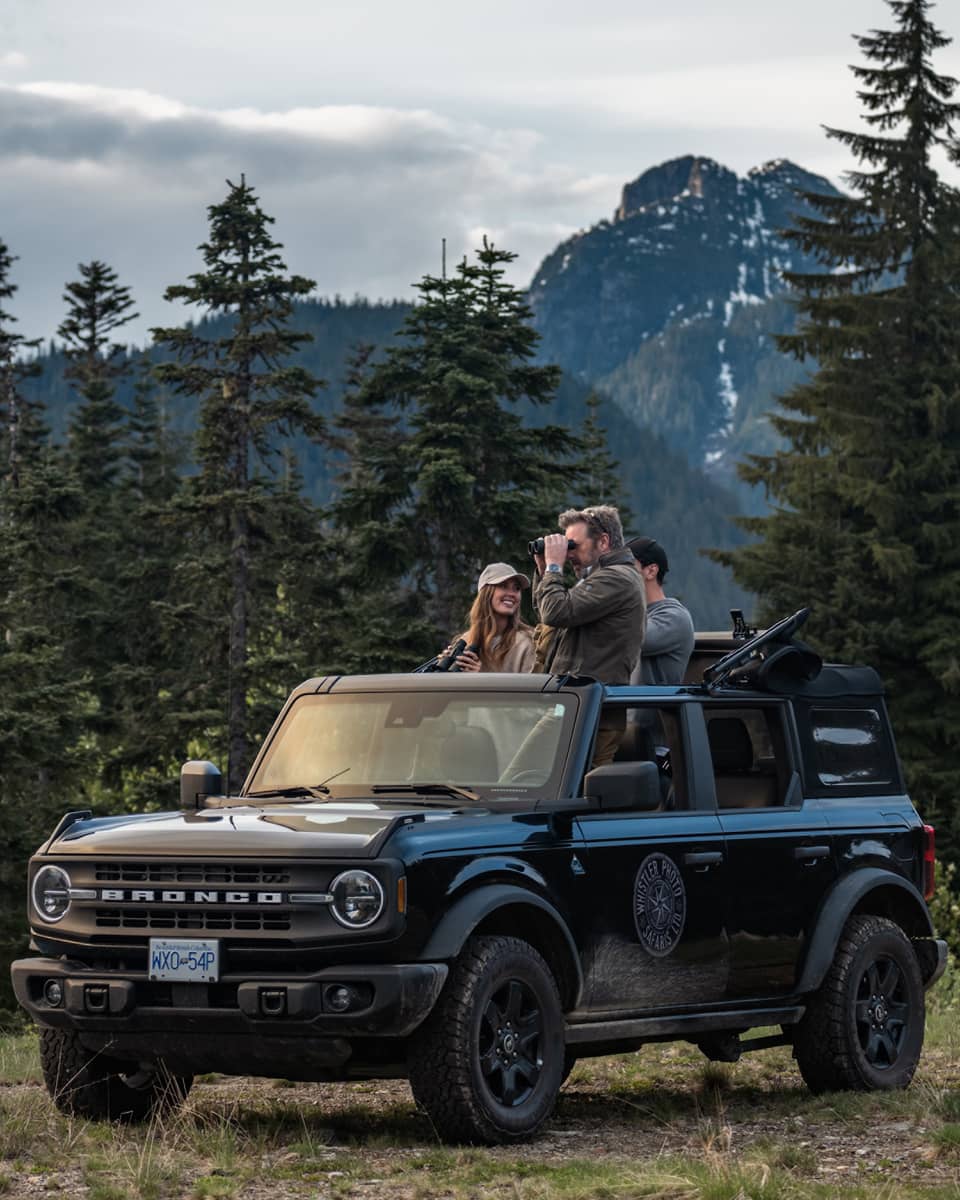 Group of friends with binoculars stand in open-top Ford Bronco surrounded by trees and mountains