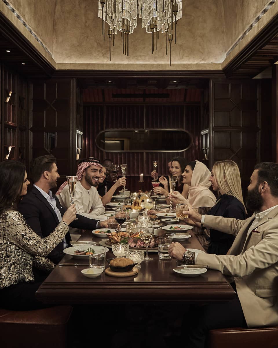 Ten people sit at a long, dark-wood rectangular table in a dark-wood panelled, speakeasy-style room with a crystal chandelier overhead and a marble bar counter and bartender to the side