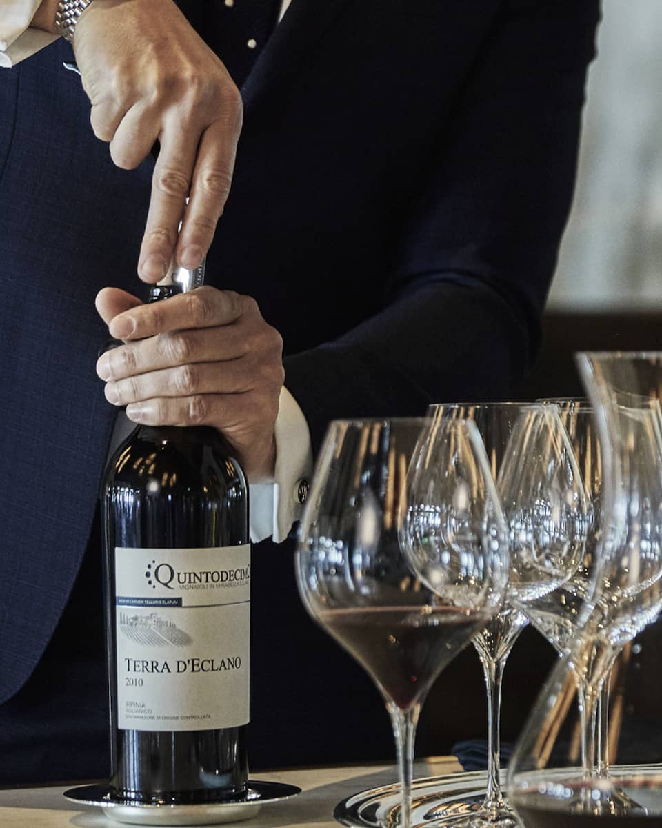 A person opening a bottle of wine, with wine glasses and additional wine bottles beside it