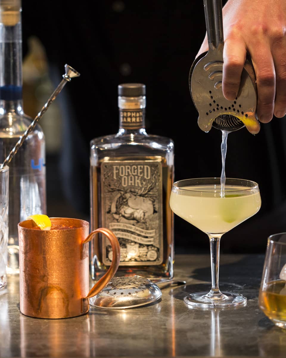 Various cocktail glasses and liquor bottles on bar as mixologist pours martini out of shaker