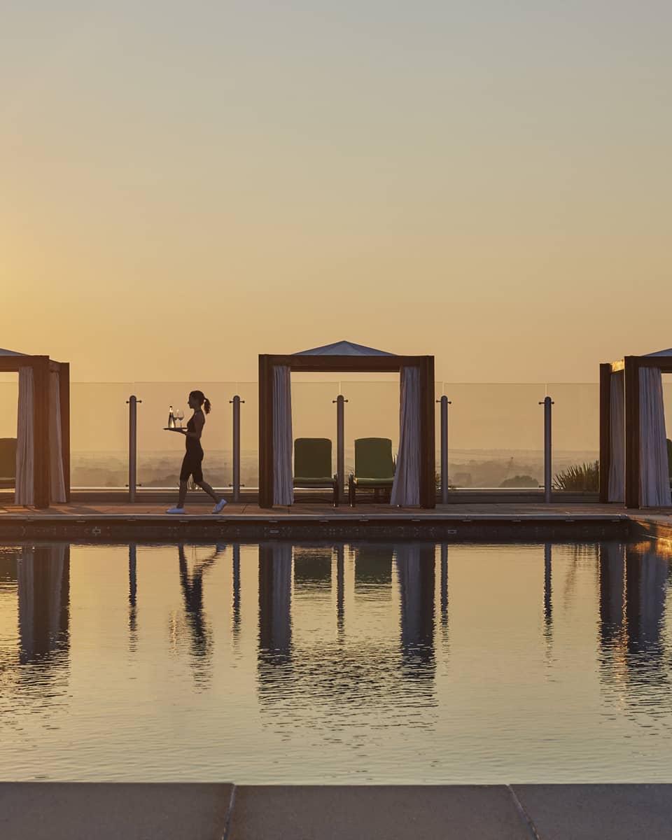 A server walking by a rooftop pool with cabanas at sunset and carrying drinks, with the golden sky reflecting on the water in a luxury hotel terrace setting