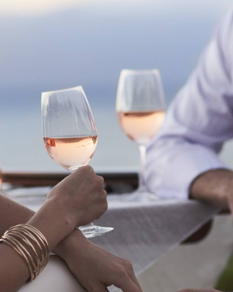 Close-up of woman and man in white, sitting and holding glasses of rose wine at dusk