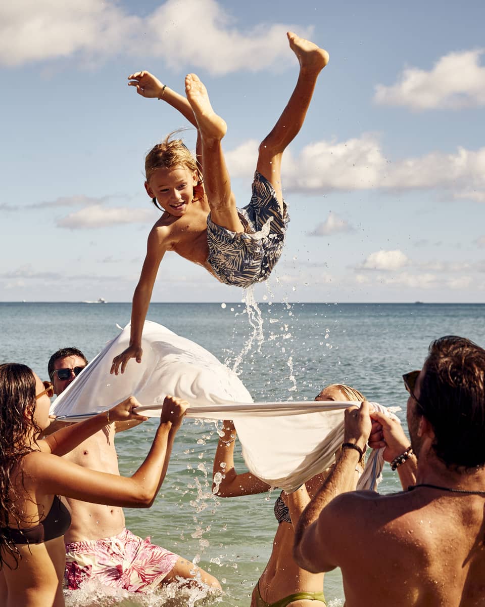 A group enjoys a sunny beach day, tossing a child into the air with a white sheet. The child smiles mid-air against clear skies.