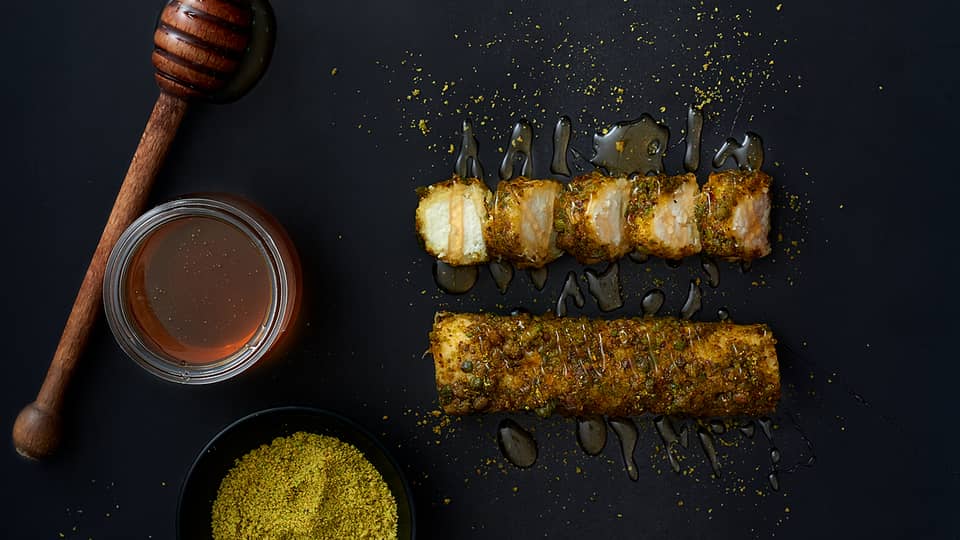 Close-up top view of black dish and item encrusted with spices and herbs, drizzled with honey, honey pot and spices beside