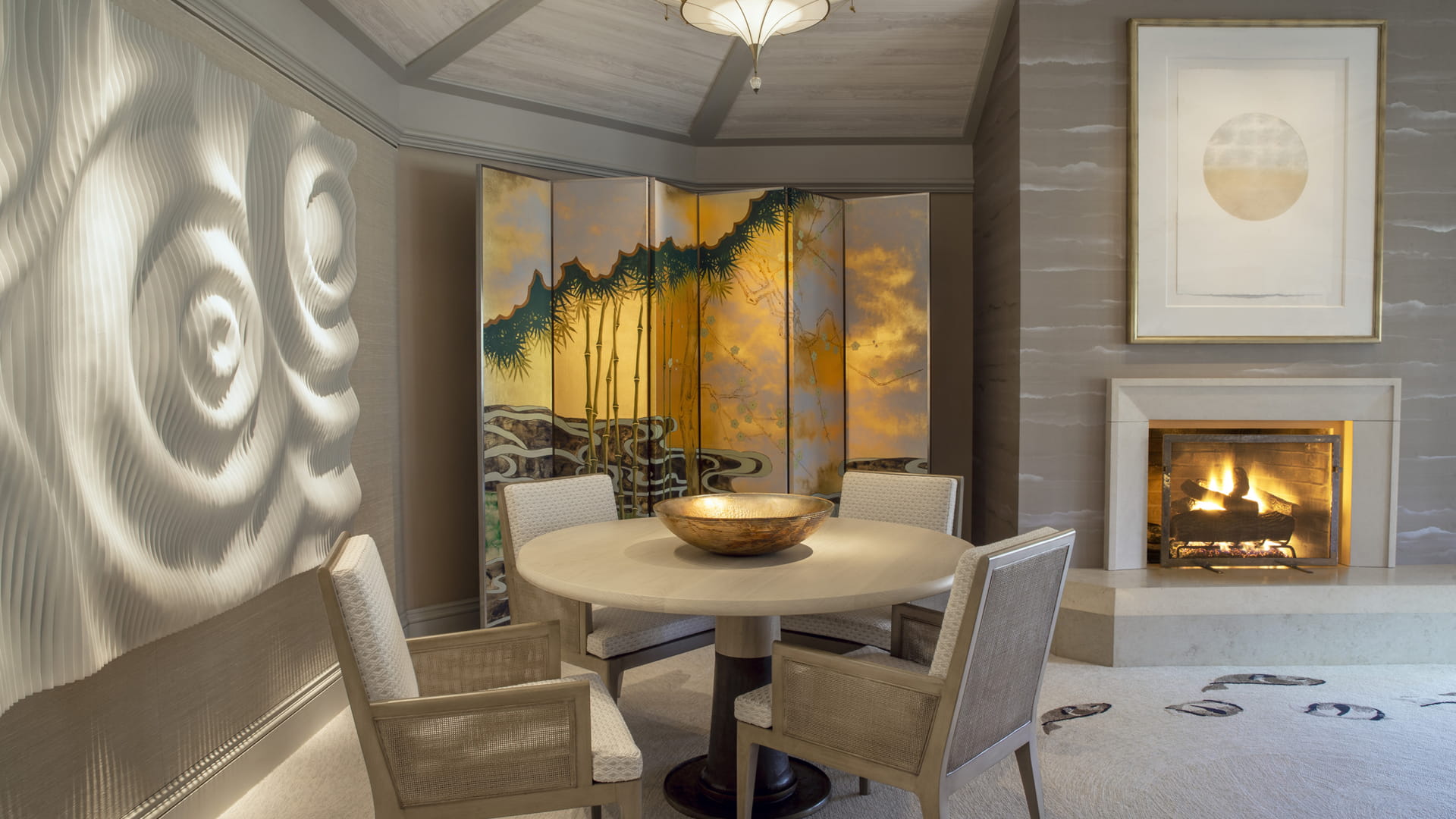 A suite sitting room where a round pale table and cane-backed chairs stand before a gold-leaf folding screen painted with pines, a carved relief wall of concentric rings on the left and a lit fire in a stone hearth on the right.