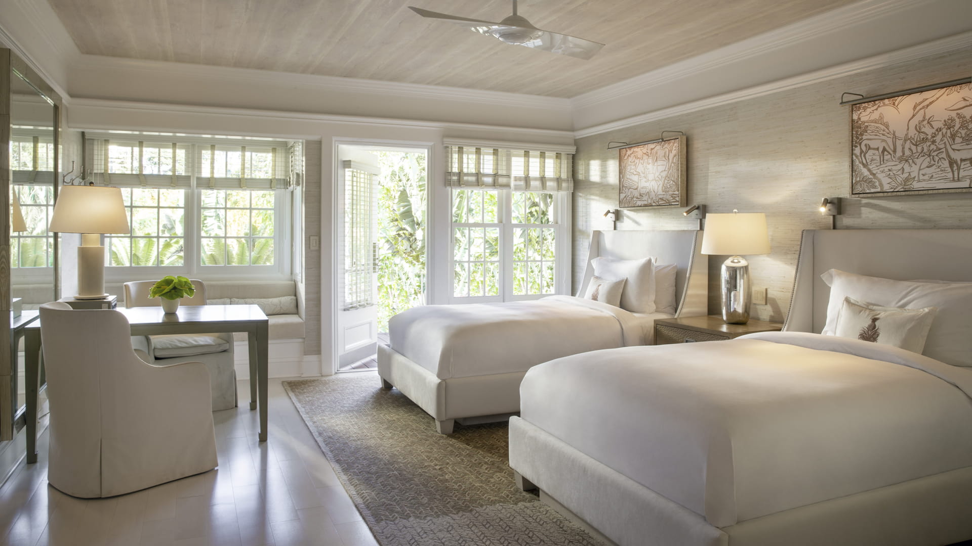 A guest room in cream and pale grey with two beds under a limed-timber ceiling and paddle fan, a window seat built into the bay of divided-light windows, a writing table and slipcovered chair, and French doors open onto green foliage.