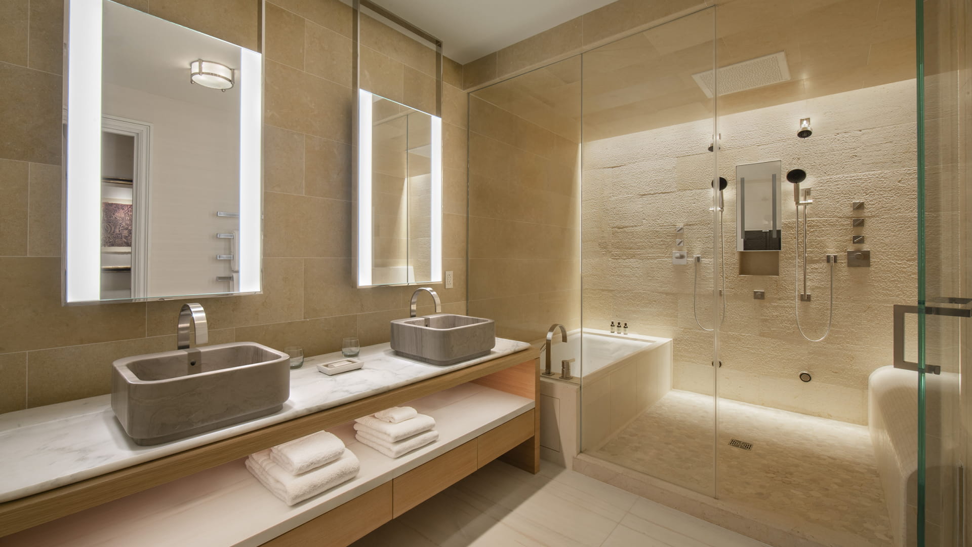 A bathroom in warm limestone with two carved stone basins on a marble and oak vanity, backlit mirrors, folded white towels on the open shelf below, and a glass-walled wet room holding a deep tub and a textured stone shower wall.