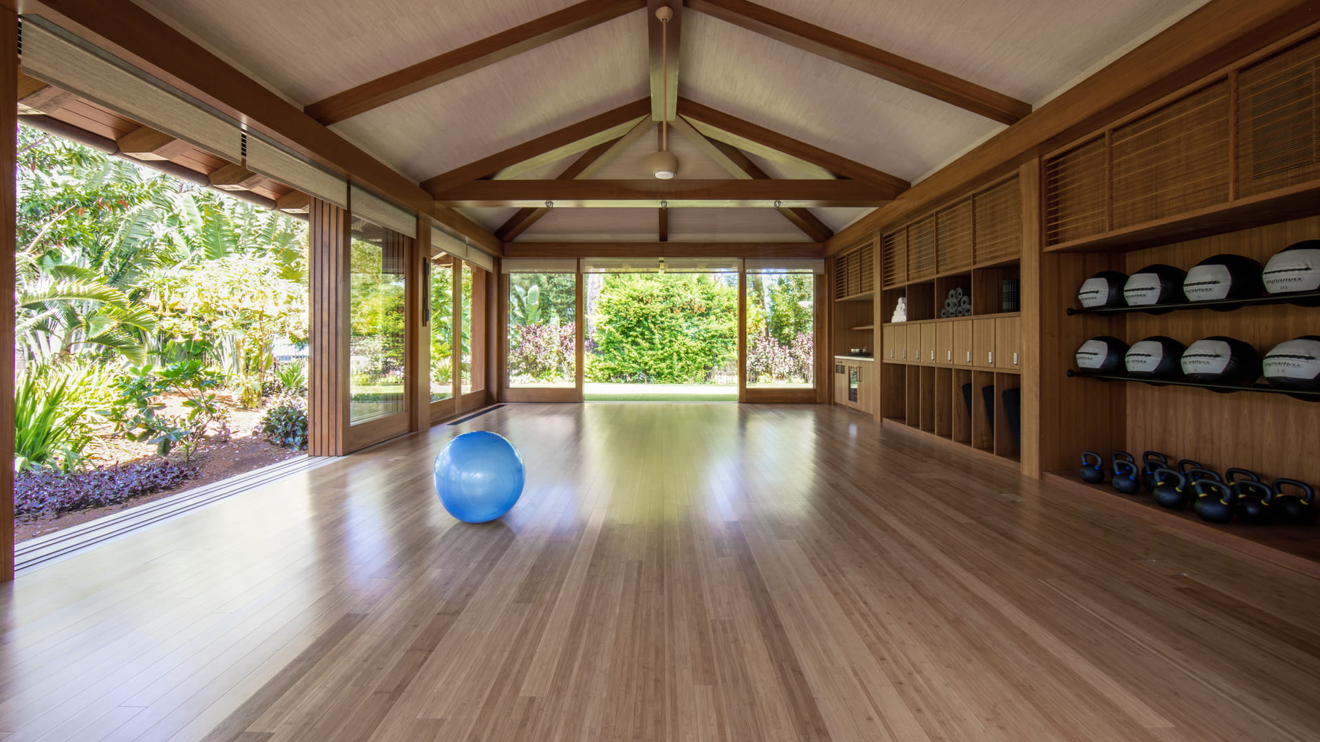 An empty movement studio under an open timber truss roof, its wide plank floor running to glass doors that slide back on two sides onto banana leaves and flowering shrubs, with kettlebells, medicine balls and louvred cabinets along the far wall.