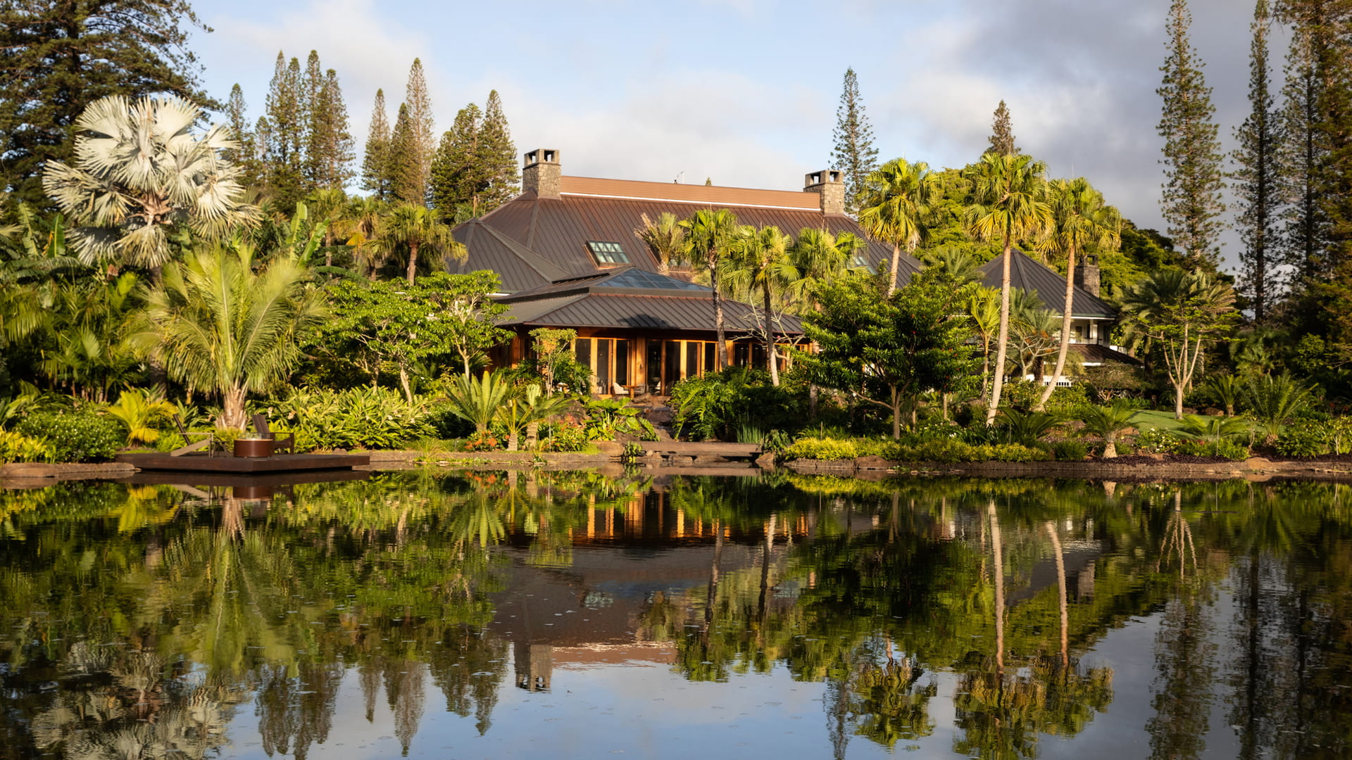 The low copper-roofed lodge seen across its still reflecting pond in early light, timber verandas and stone chimneys half hidden by fan palms and clipped shrubs, with tall Cook pines ranged along the ridge behind and the whole scene doubled in the water.