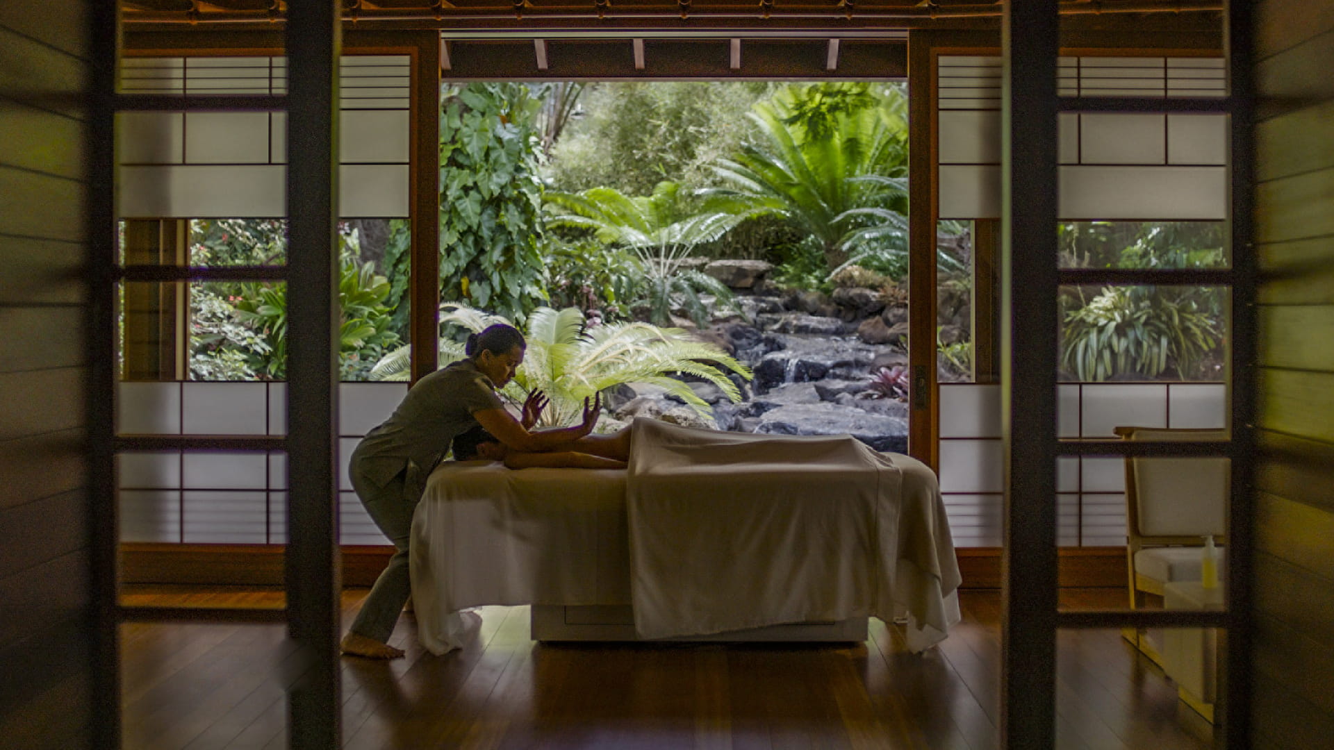 A treatment hale seen through its dark timber frame and shoji screens, a therapist working at a draped table on a polished wooden floor while the far wall stands fully open to a rock-lined stream and a bank of ferns and cycads.