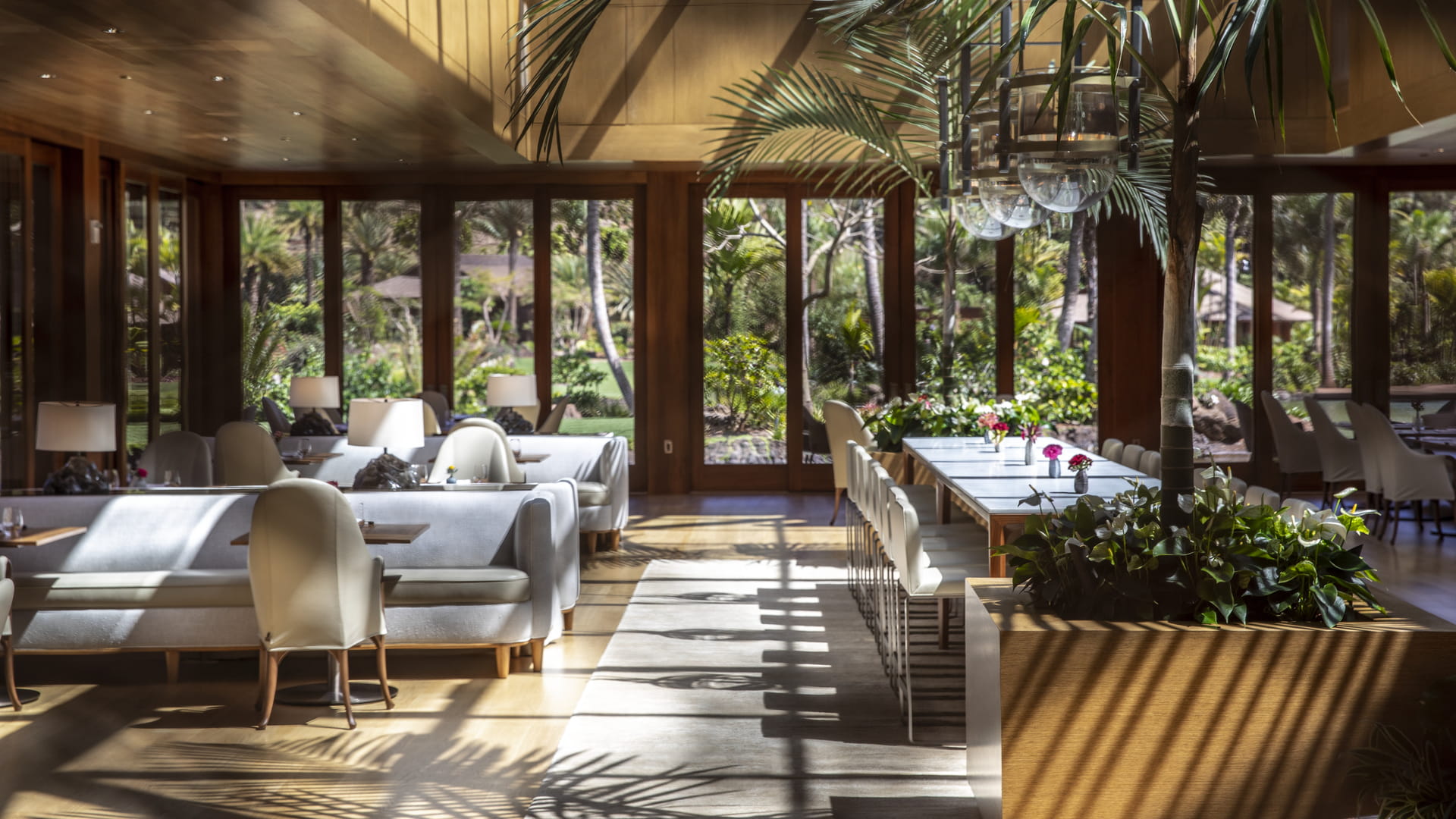 A dining room panelled in dark koa-toned timber with full-height glazing on every side, palm fronds reaching in over a long banquet table, upholstered banquettes and cane-back armchairs, and hard morning shadows striping the pale floor.
