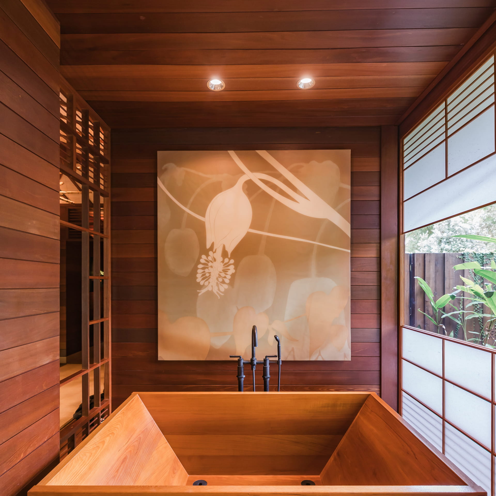 A deep square soaking tub cut from pale cedar in a room lined entirely in red-brown timber boards, a large soft-focus painting of orchid stems on the wall above the black tub filler and a shoji-screened window onto planting outside.
