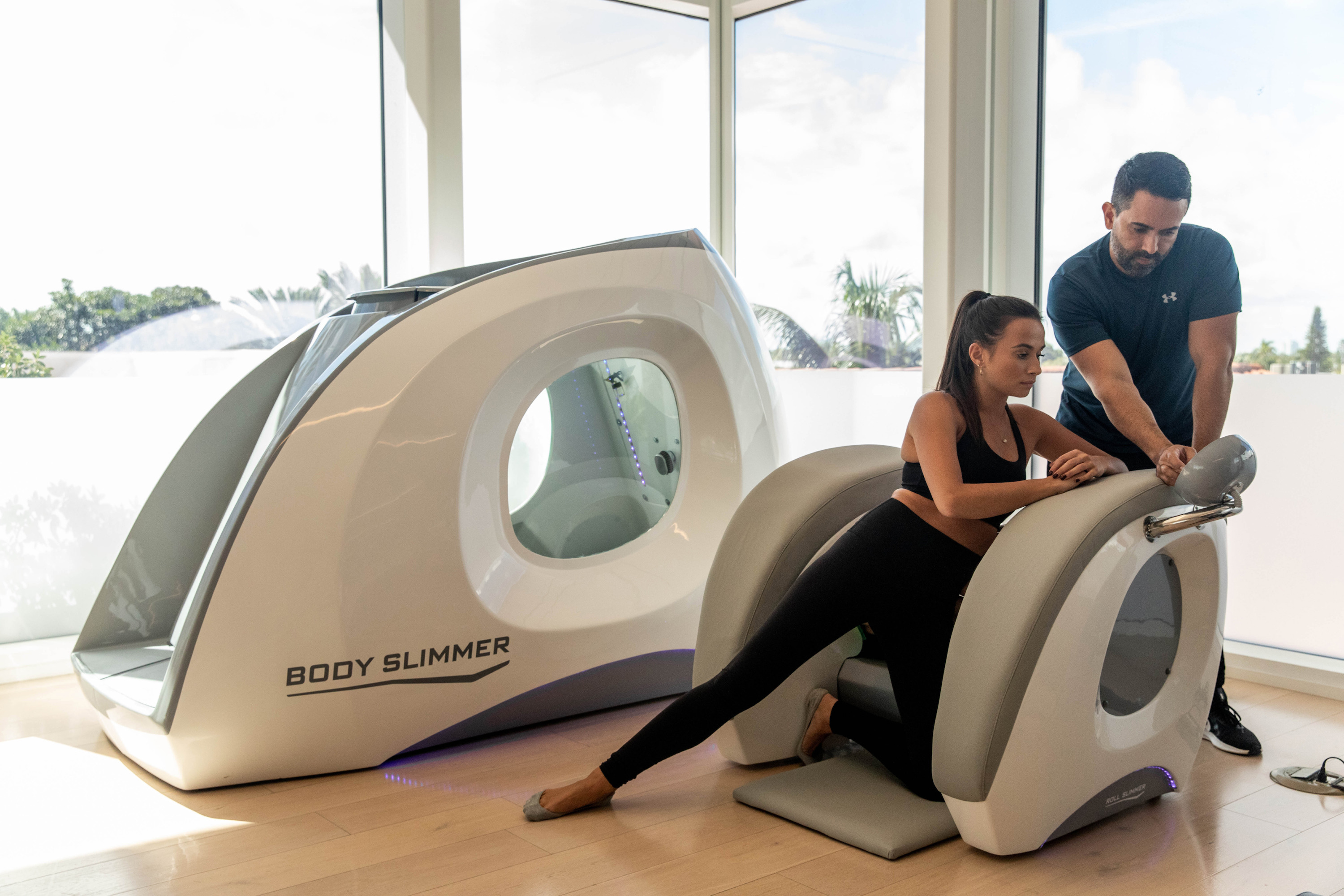 A woman in black sportswear leans into a machine labelled BODY SLIMMER while a man steadies her.