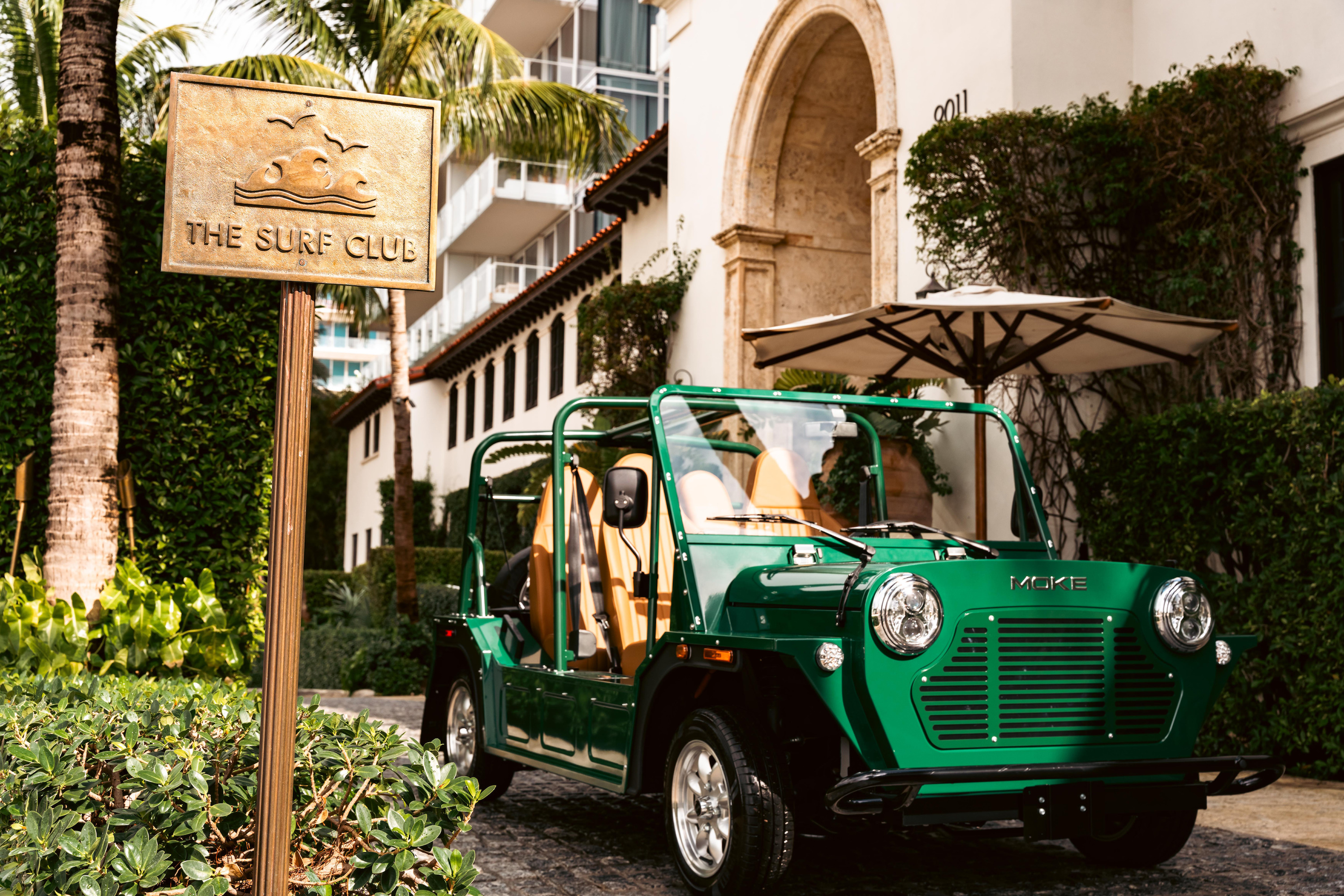 A green open-sided electric Moke parks beside a brass sign reading THE SURF CLUB.