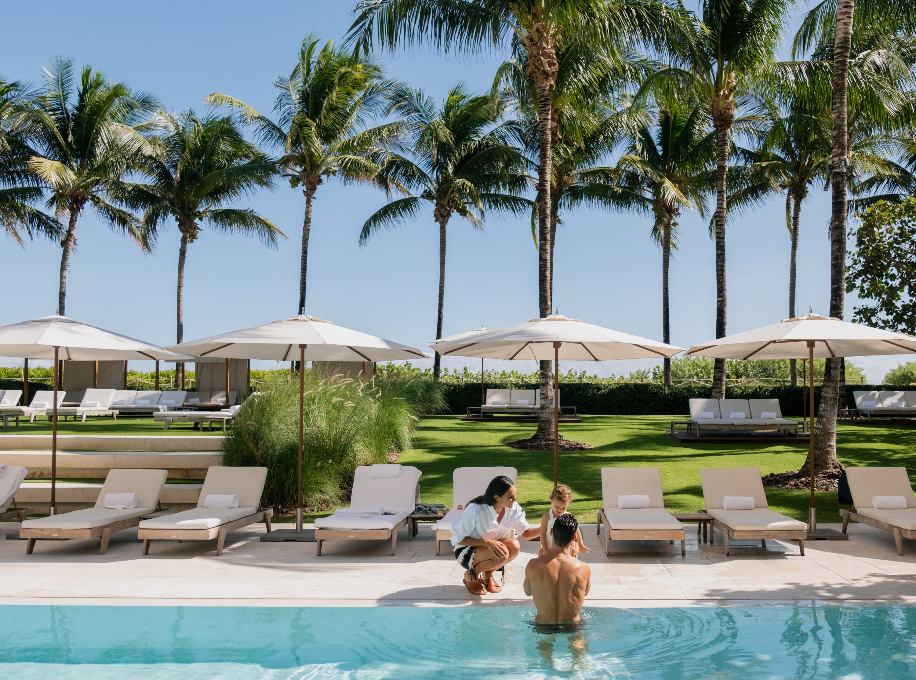 White loungers and parasols sit on a pool deck as an adult and a child stand at the water's edge.