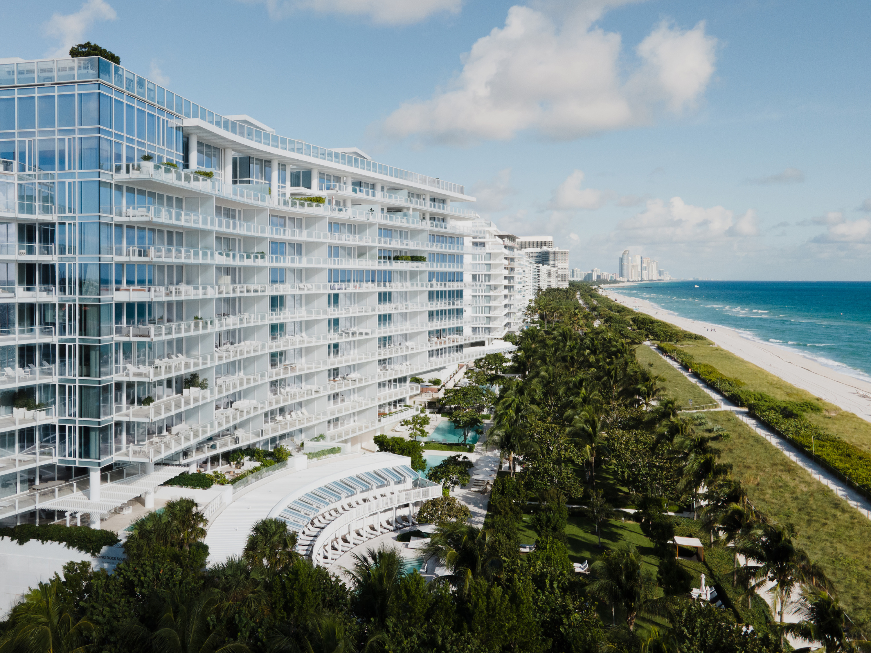 From above, a tall white glass building stands beside a long sandy beach and blue sea.