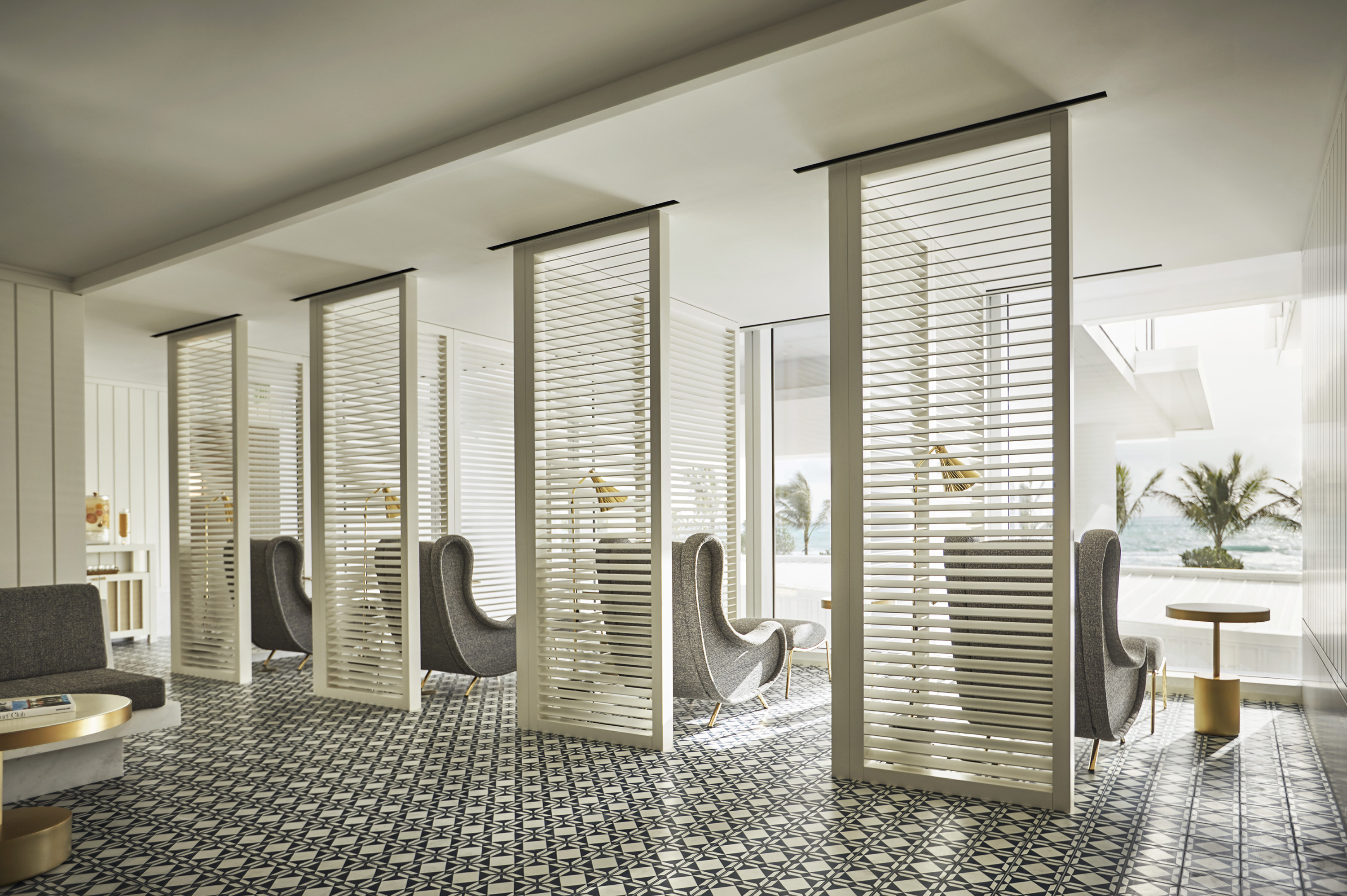 A brass low table sits between winged armchairs in a room with white slatted screens and palms outside a window.