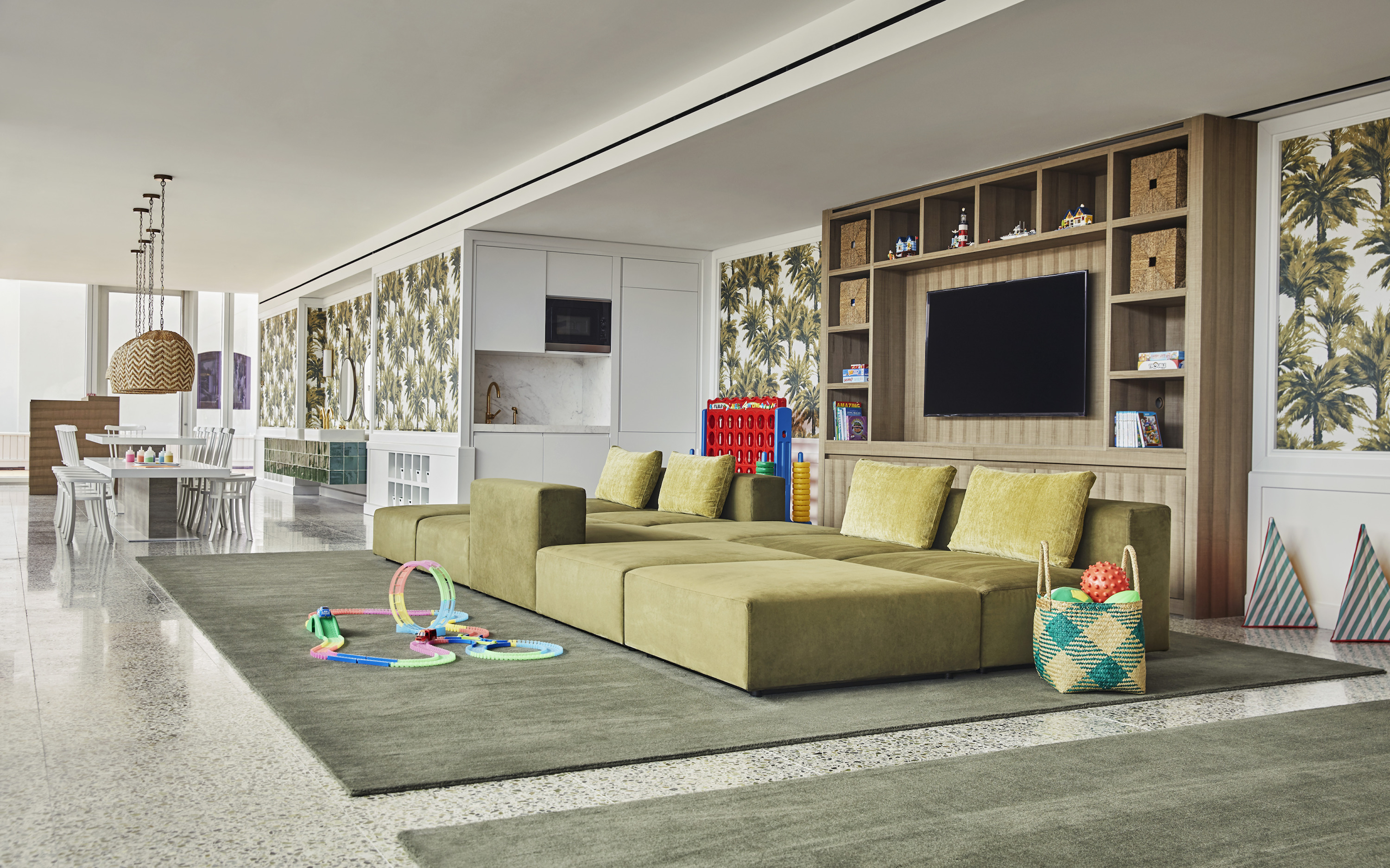 A long olive-green sofa faces a television in a lounge with palm-print wallpaper and children's games.