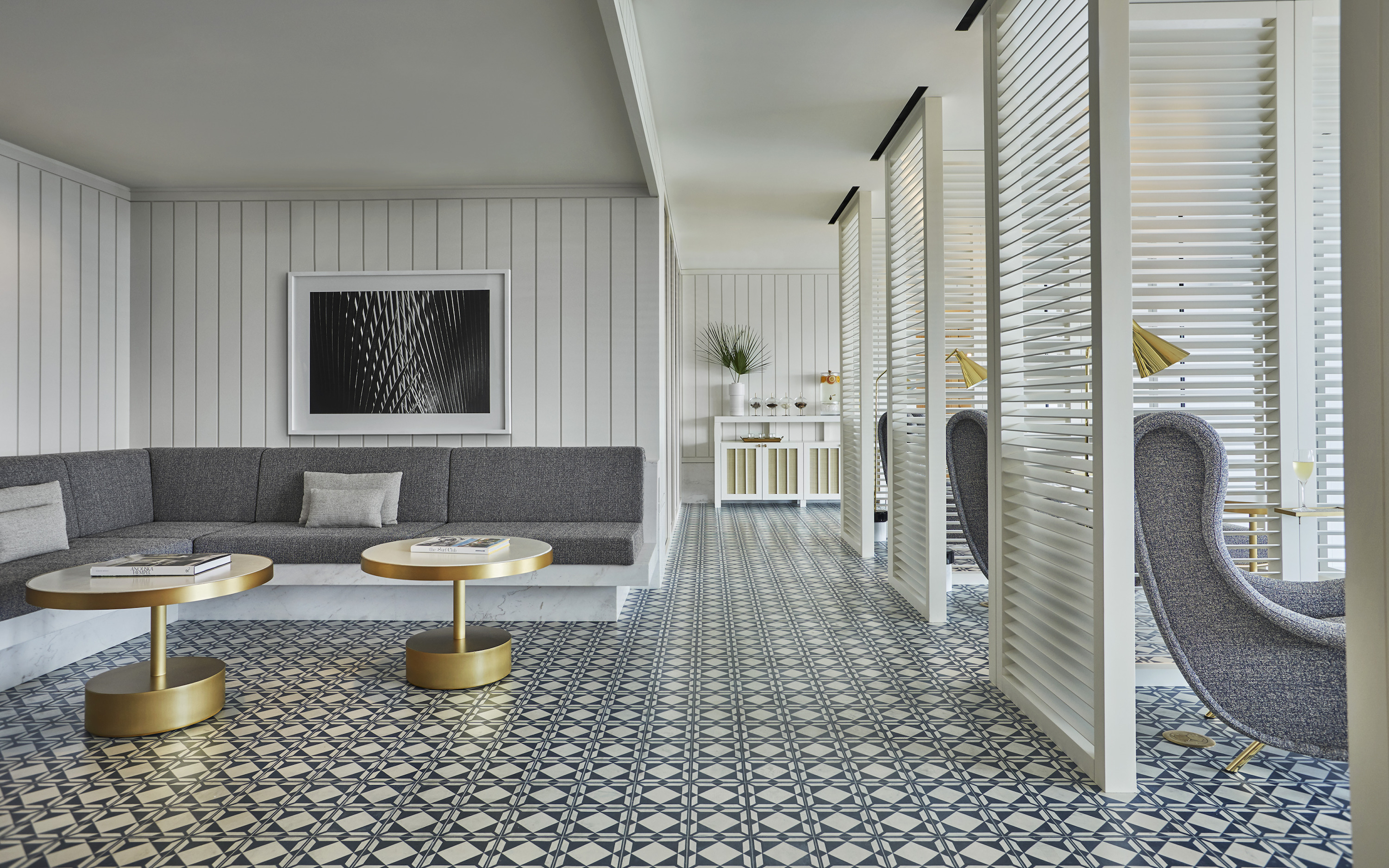 A built-in sofa and two round gold-edged tables sit beside white slatted screens under a framed black and white abstract.