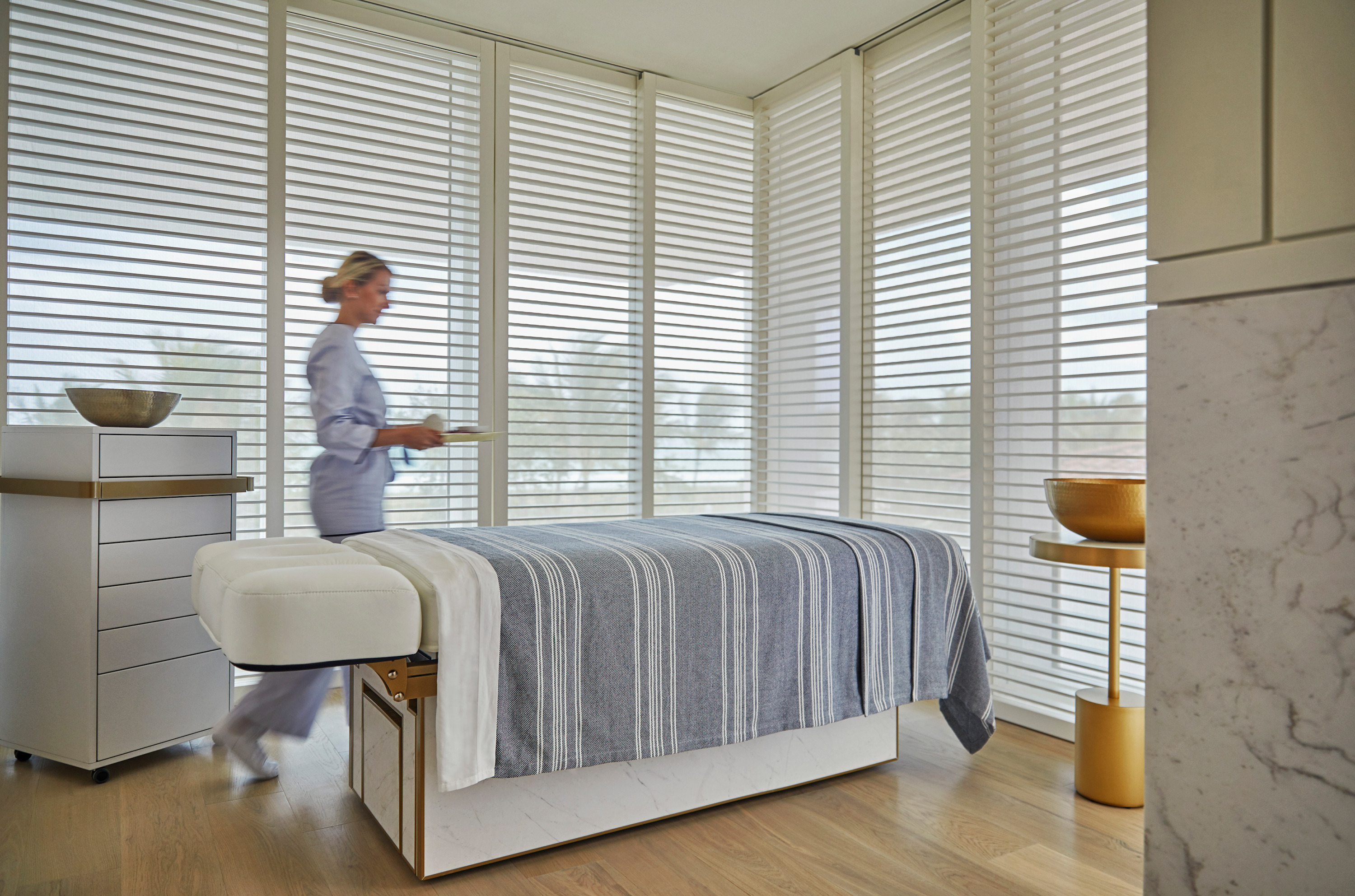 A therapist in pale blue walks past a massage table in a corner room with slatted shutters and wood floor.
