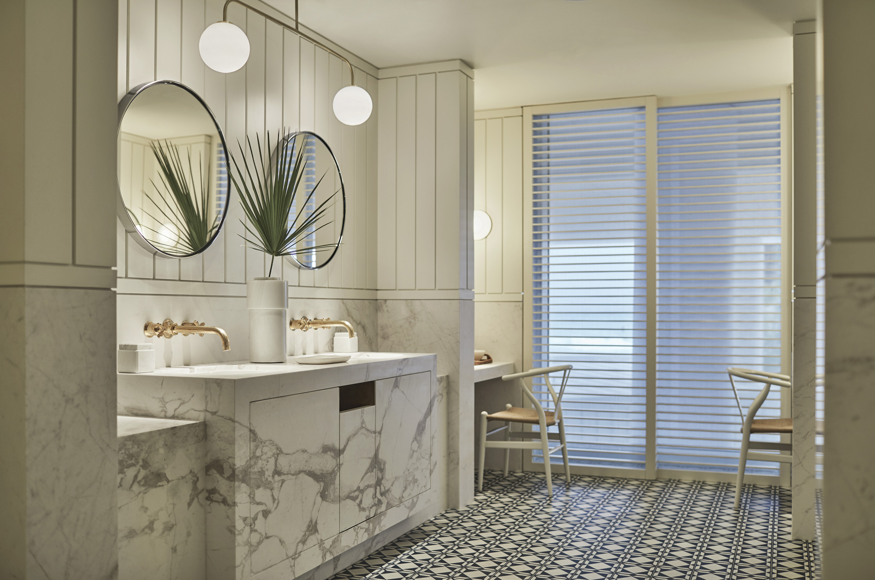 A marble vanity with two round mirrors and a vase of palm fronds faces slatted blinds over a tall window.