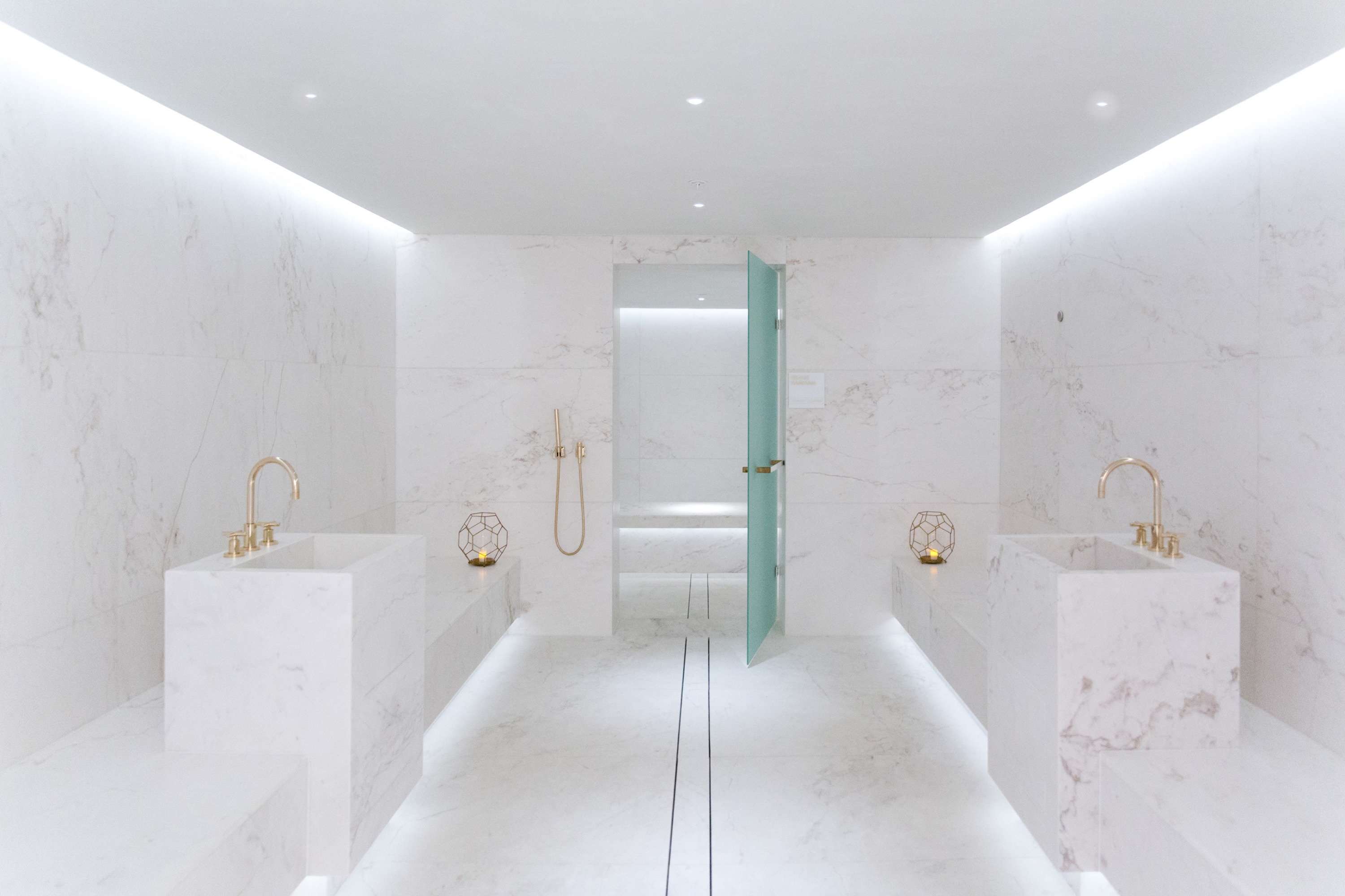 Two block basins with brass taps sit in a white marble room with a hand shower and pale-green frosted door.