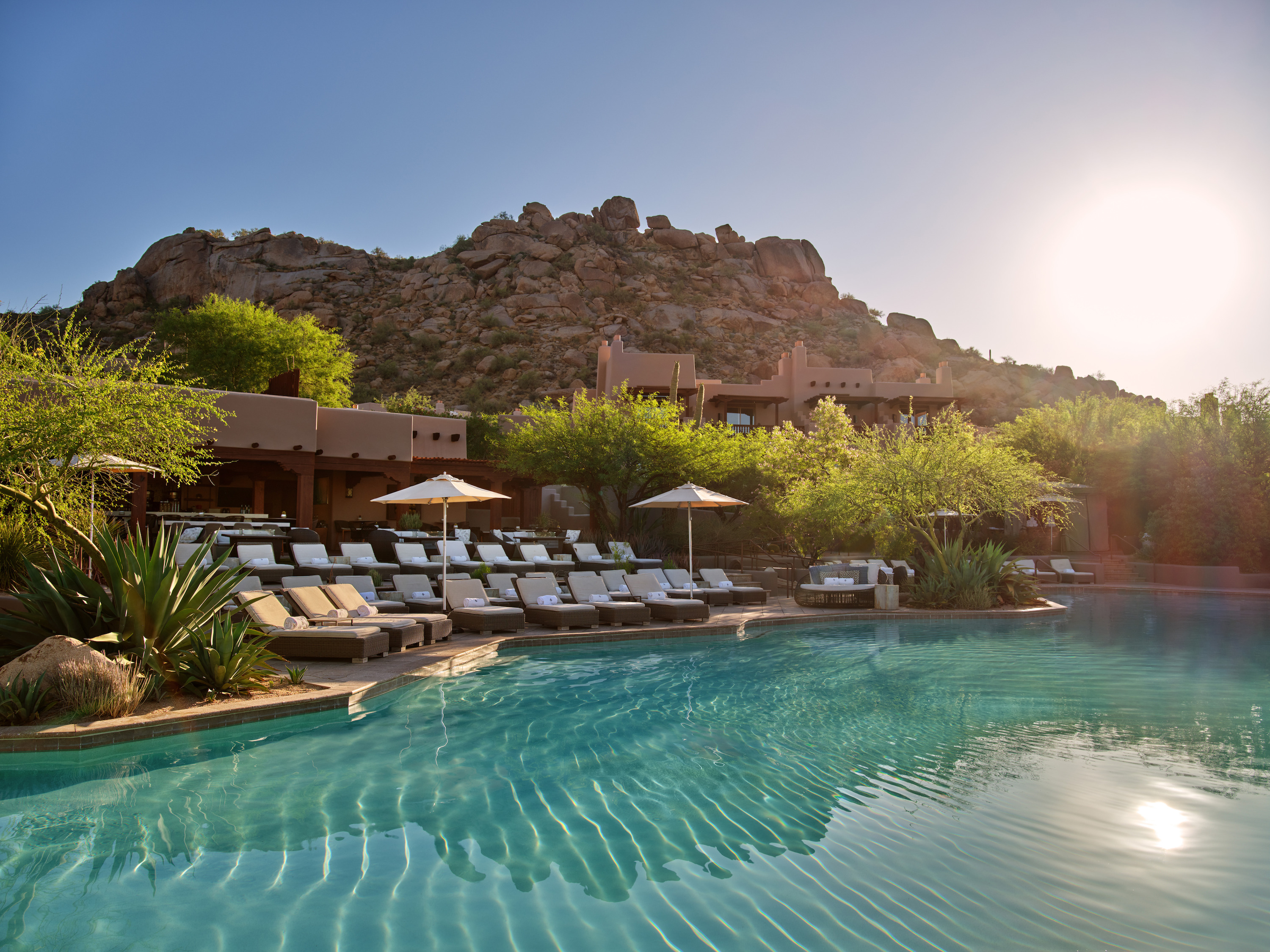 Ripples cross a bright pool in low sun beside rows of loungers, a few umbrellas, and tan buildings, with a boulder hill behind.