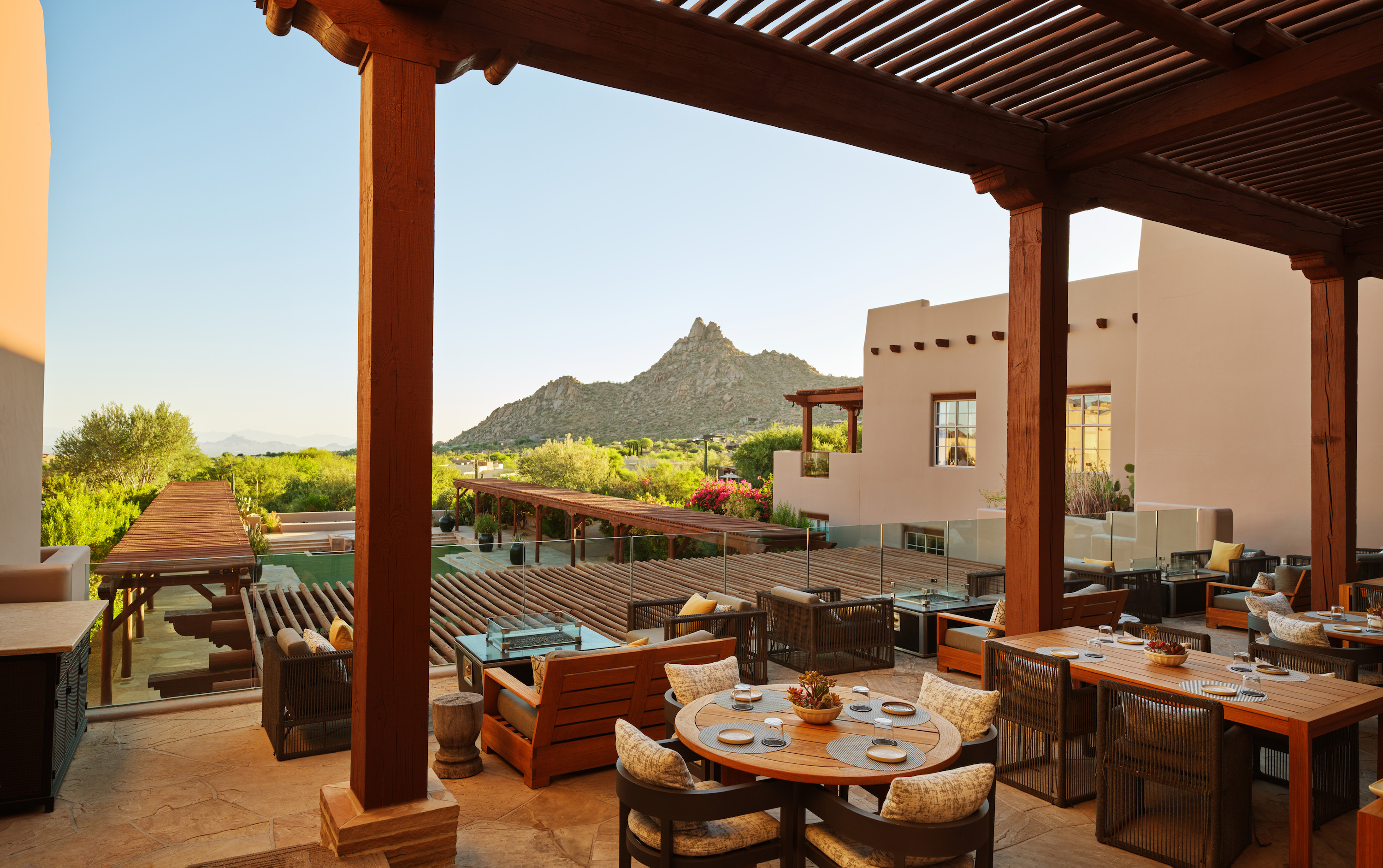 A dining terrace under a timber pergola has set tables with rattan chairs, a stone building, and a pointed mountain.