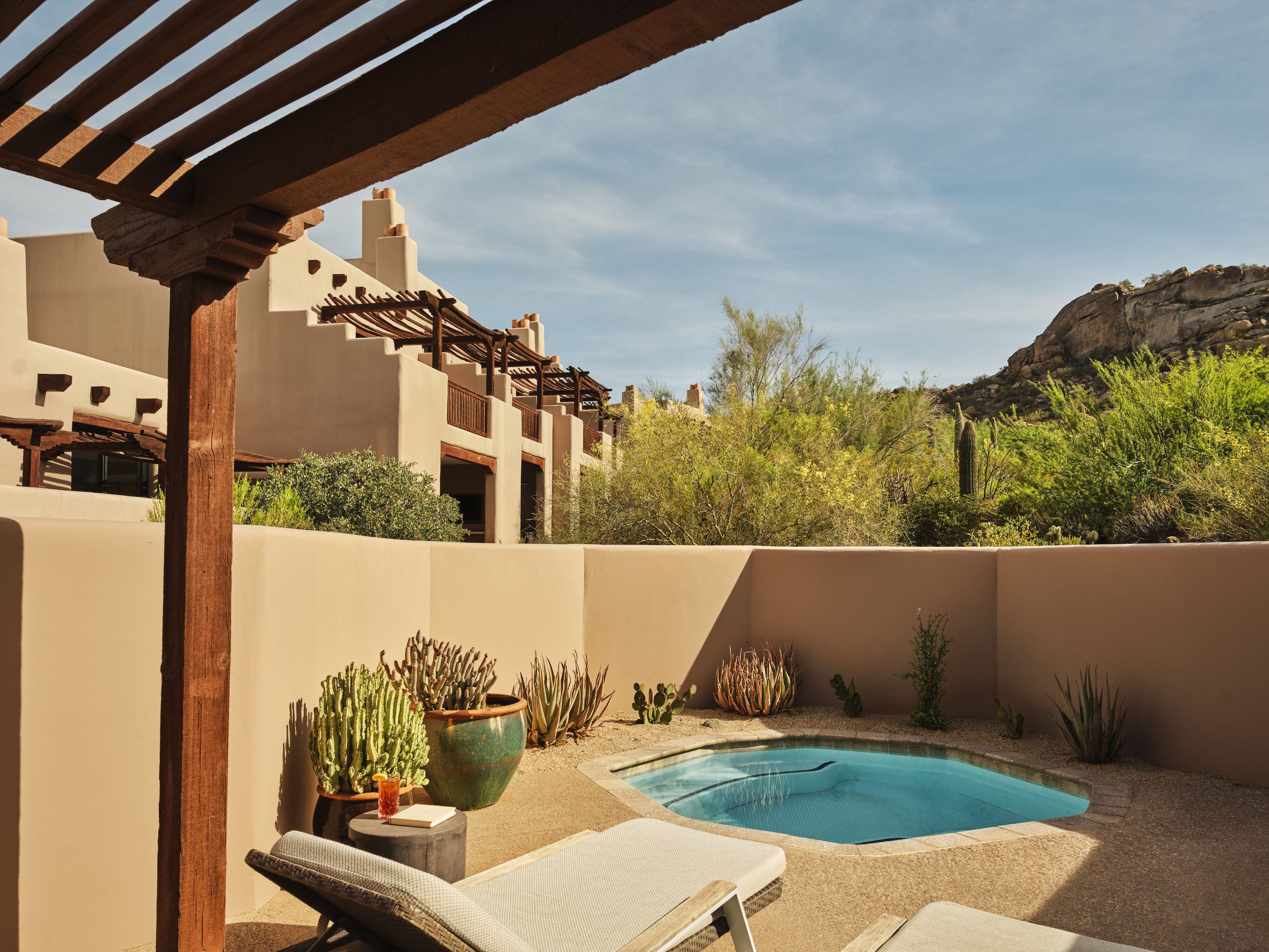 A walled desert patio has a small blue pool, a lounger, potted cactus, and tan buildings under a rocky hill.