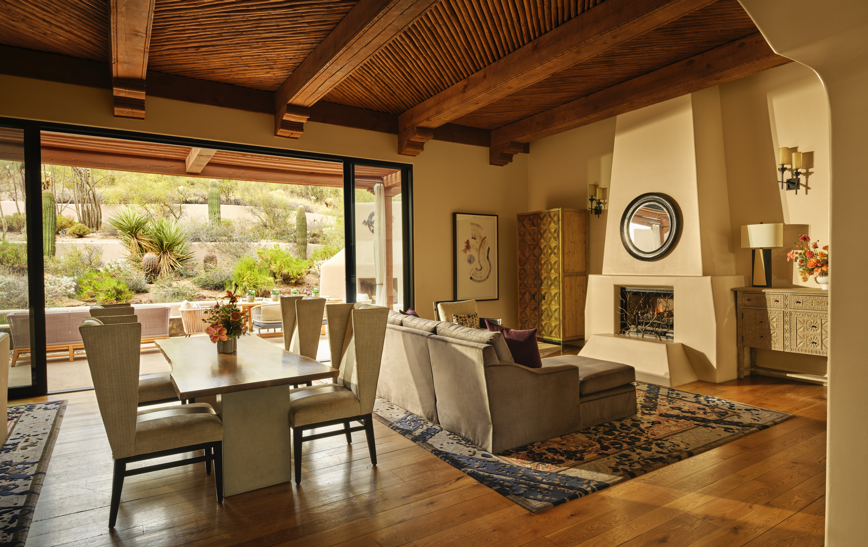 A long wooden dining table with tall-backed chairs, a pale fireplace with a round mirror, and glass doors to a desert patio are in the same room.