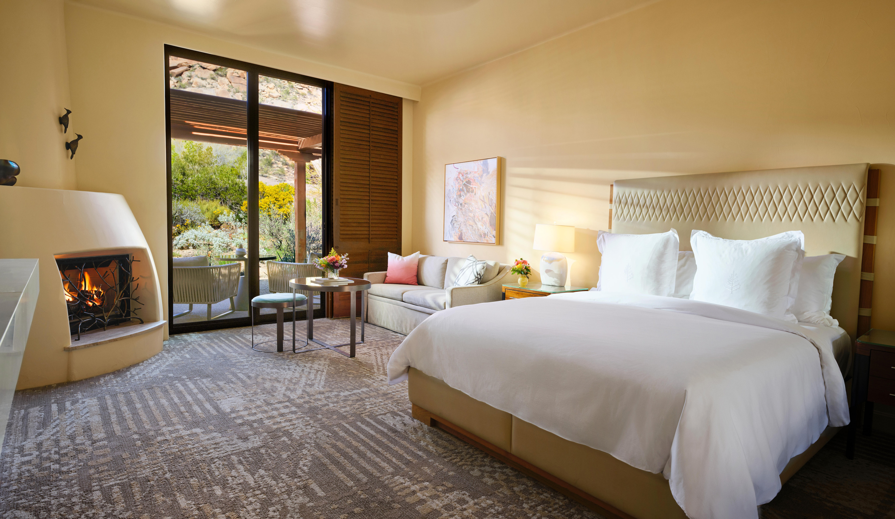 A large bed with white linens and a woven headboard, a round fireplace, and glass doors are in a room with grey patterned carpet.