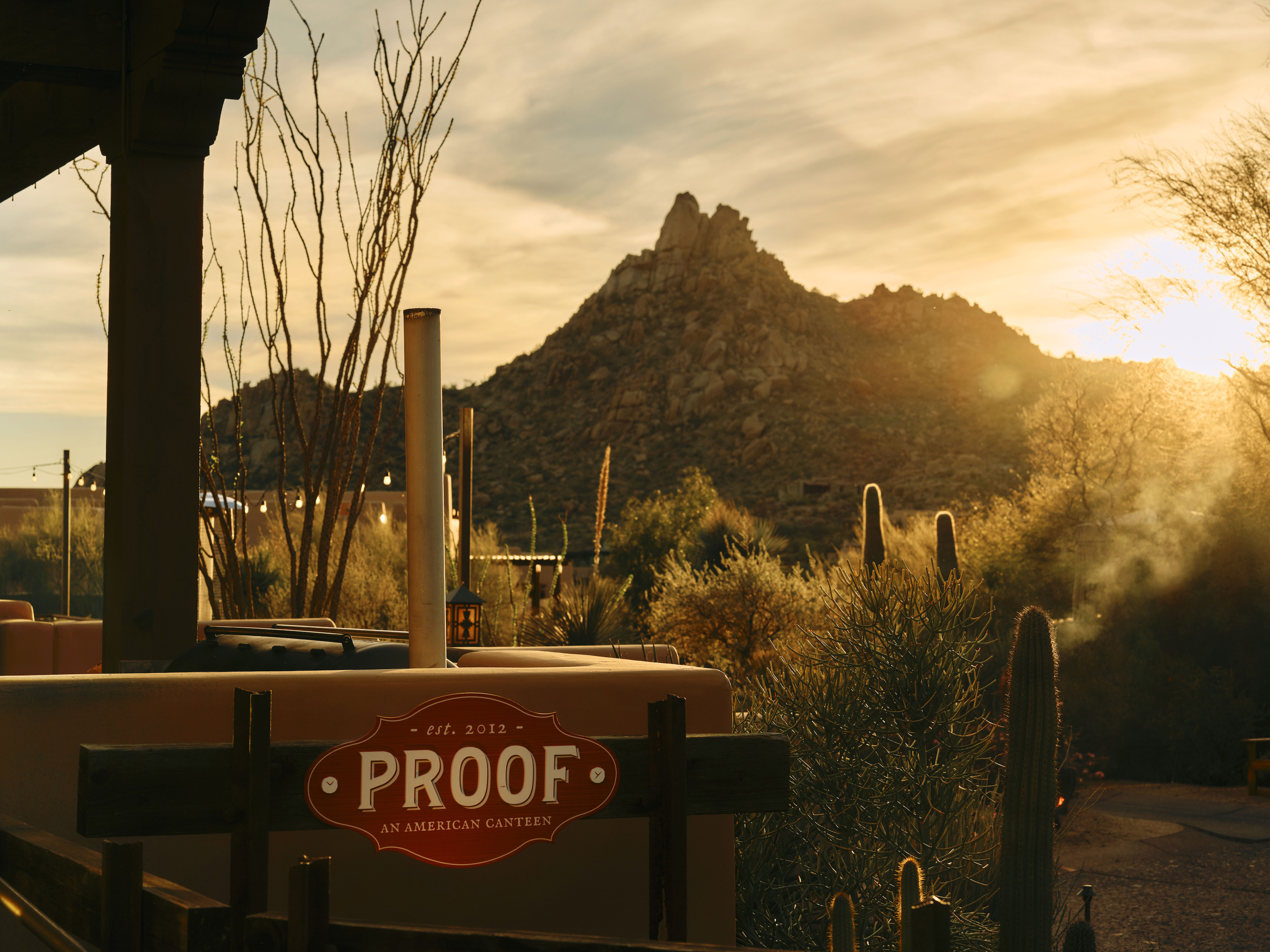 A red round wooden PROOF sign stands between dry shrubs and a tall cactus, with a rocky peak and sun flare behind.
