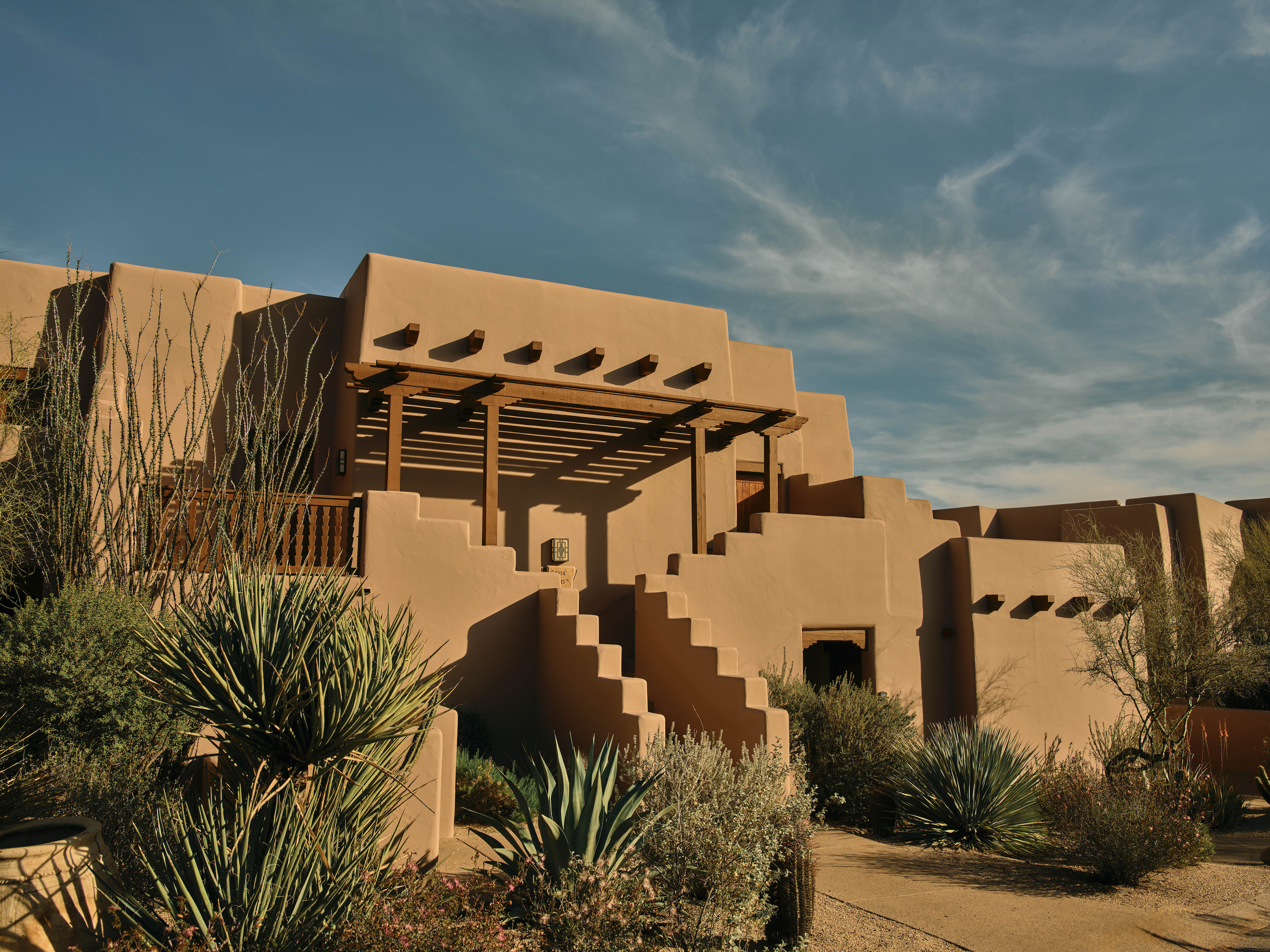 A stepped tan building of two and three stories with flat roofs and a wooden pergola sits among agave and yucca under blue sky.