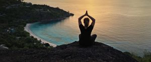 A woman sits a top of peak at sunset overlooking the ocean