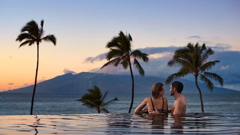 A man and a woman look at each over in an infinity pool overlooking the ocean