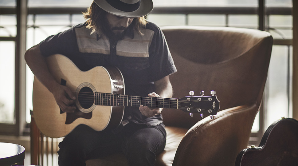Man in a wide-brim hat plays the guitar in a brown chair