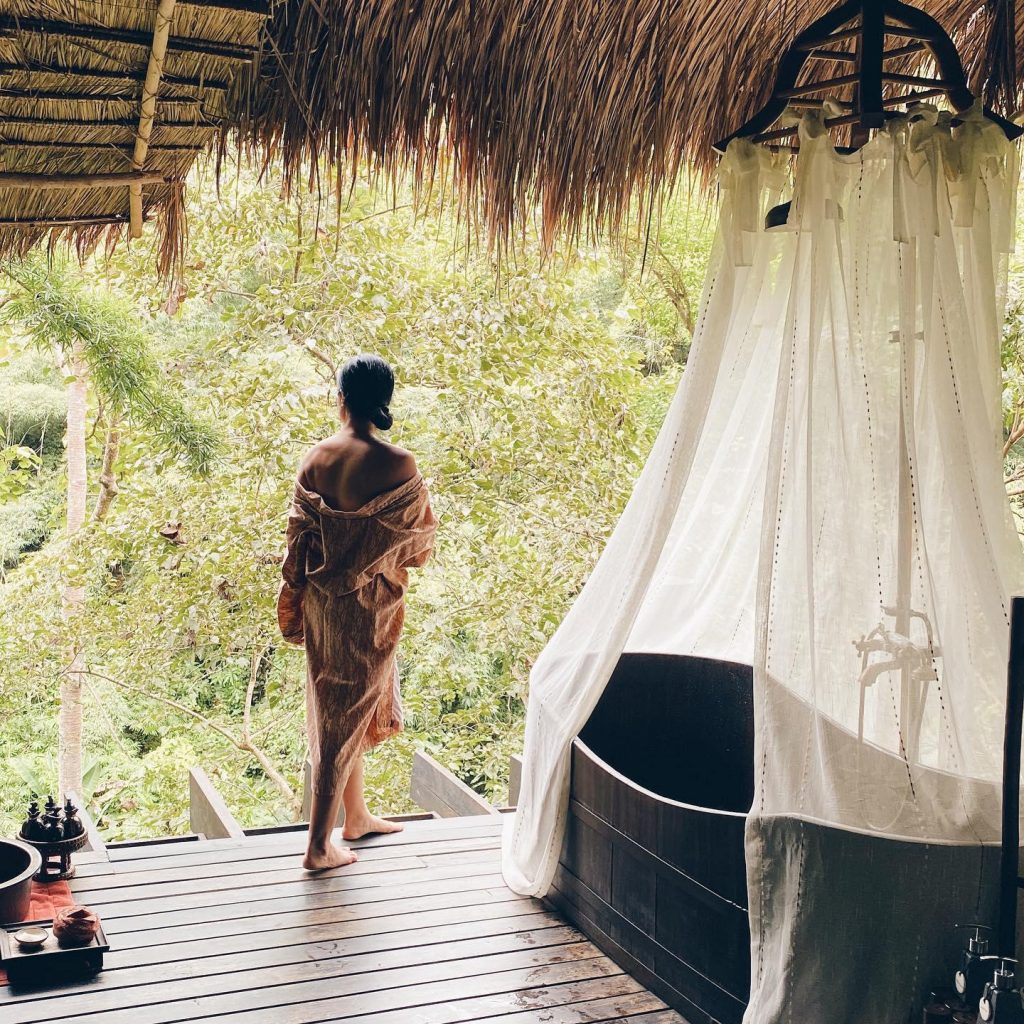A woman stands on a deck in a floral robe next to a canopied bathtub
