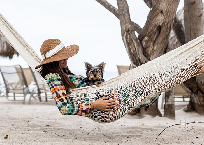 A woman in a straw hat sits in a hammock on the beach with a small dog in her lap