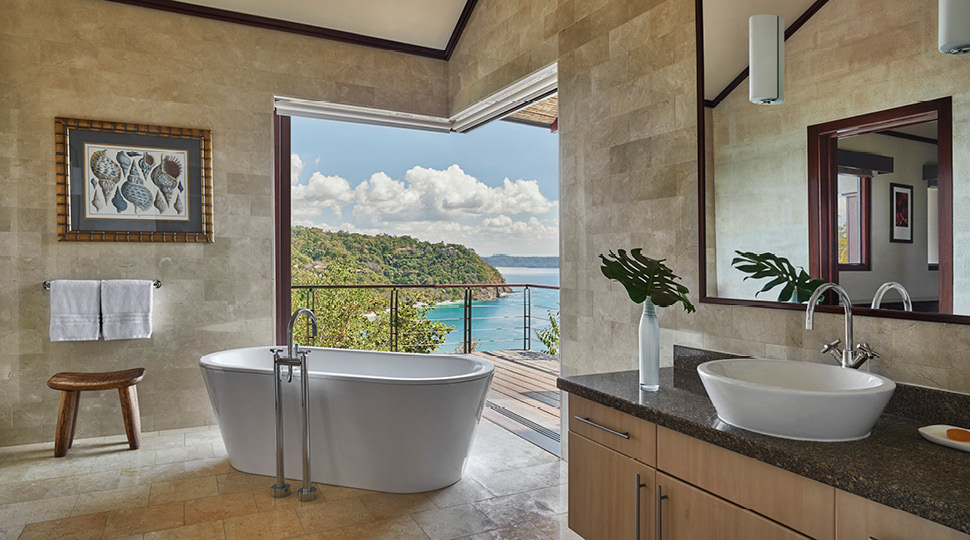 Bathroom with a vanity, mirror and a free-standing white bathtub opens up to an outdoor terrace with views of the ocean
