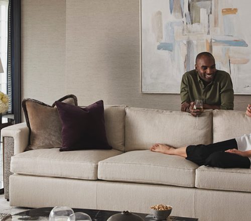 A woman lounges on a tan couch as a man leans on the back