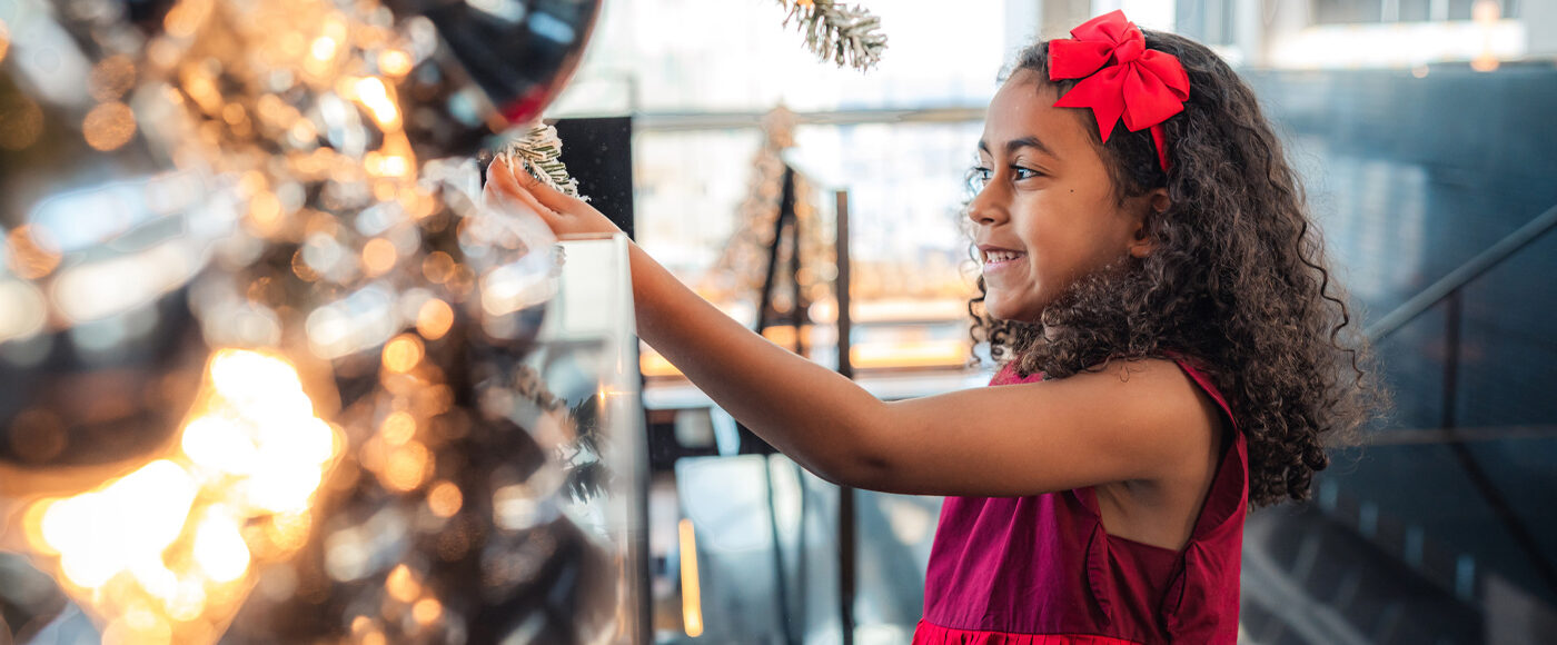 A young girl in a red dress touches the branch of a festive tree